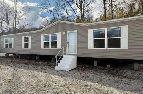 A beige, single-wide mobile home with white shutters and door stands on gravel. Bare trees are in the background under a partly cloudy sky.