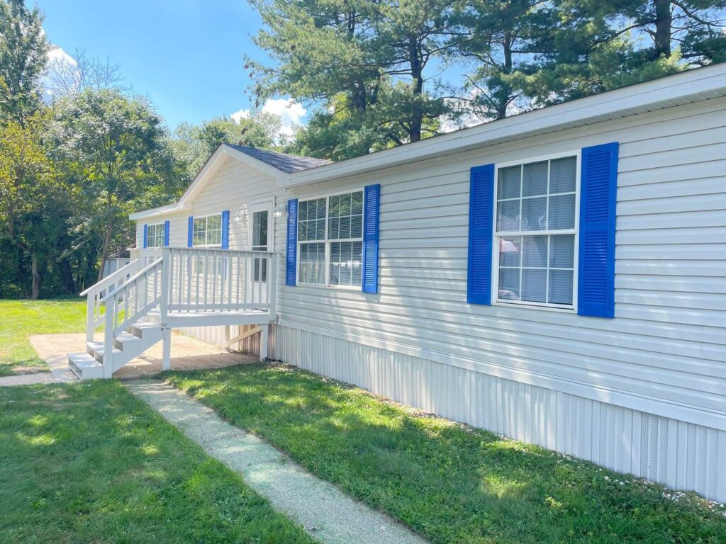 A white mobile home with blue shutters and a small porch is surrounded by lush green grass and trees. It's a sunny day with a clear blue sky.