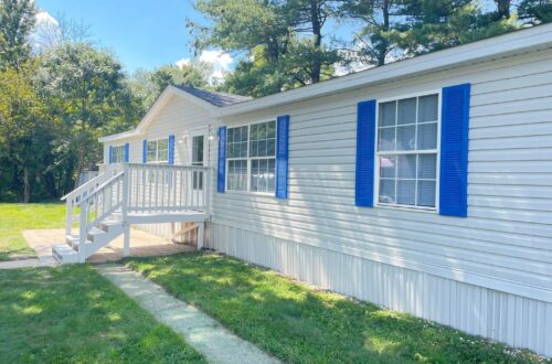 A white mobile home with blue shutters and a small porch is surrounded by lush green grass and trees. It's a sunny day with a clear blue sky.