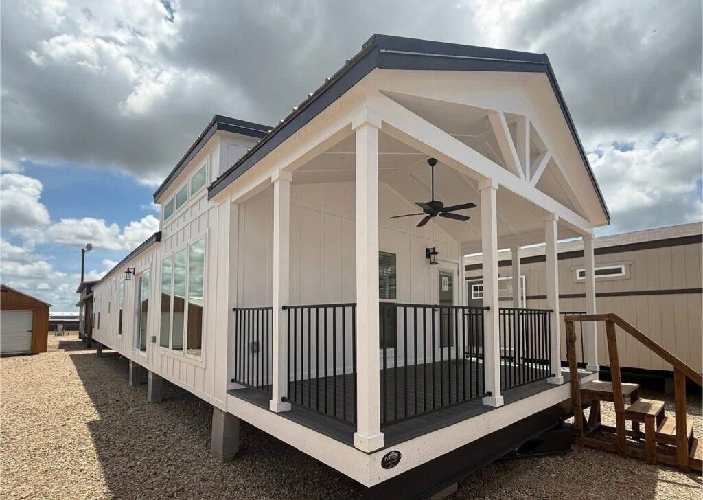 Modern tiny house with a covered porch, black railing, and ceiling fan. White exterior, large windows, set against a cloudy sky and gravel ground.
