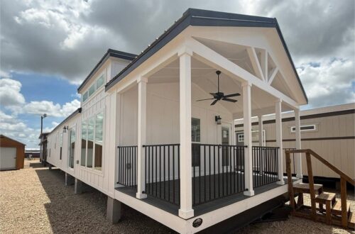 Modern tiny house with a covered porch, black railing, and ceiling fan. White exterior, large windows, set against a cloudy sky and gravel ground.