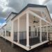 Modern tiny house with a covered porch, black railing, and ceiling fan. White exterior, large windows, set against a cloudy sky and gravel ground.