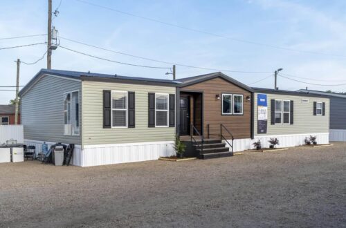 A modern manufactured home with beige and brown siding, black shutters, and steps leads to the entrance. It's set on a gravel lot under a clear sky.