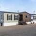 A modern manufactured home with beige and brown siding, black shutters, and steps leads to the entrance. It's set on a gravel lot under a clear sky.