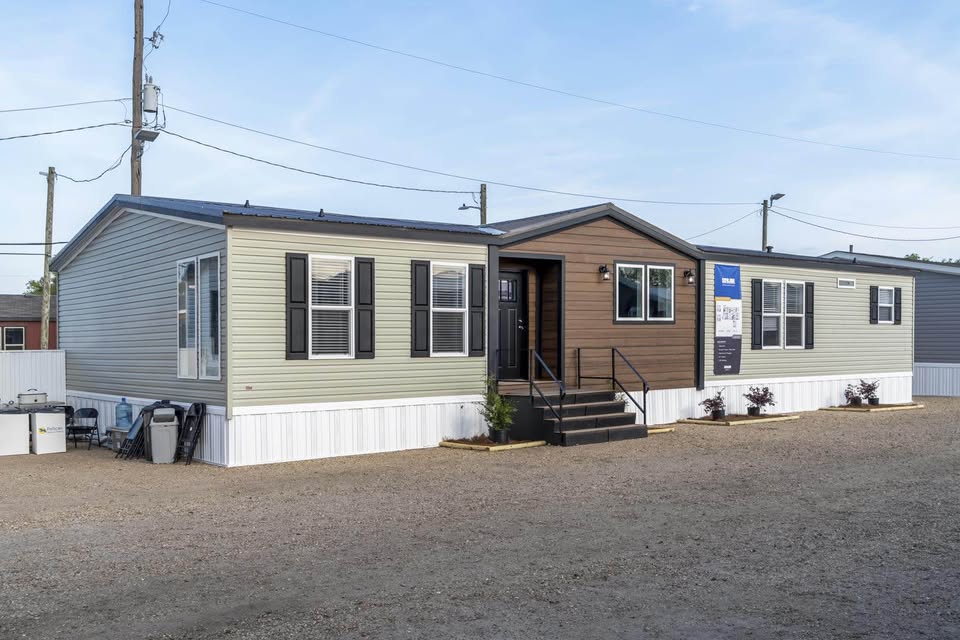 A modern manufactured home with beige and brown siding, black shutters, and steps leads to the entrance. It's set on a gravel lot under a clear sky.