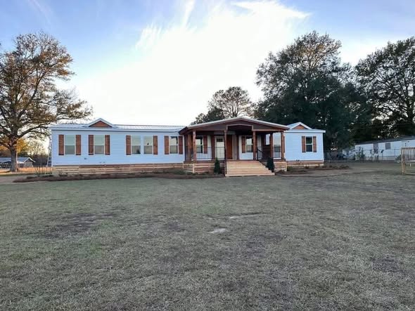 A single-story white house with a wooden porch and steps stands amid an open grassy yard, bordered by tall trees under a bright, clear sky.
