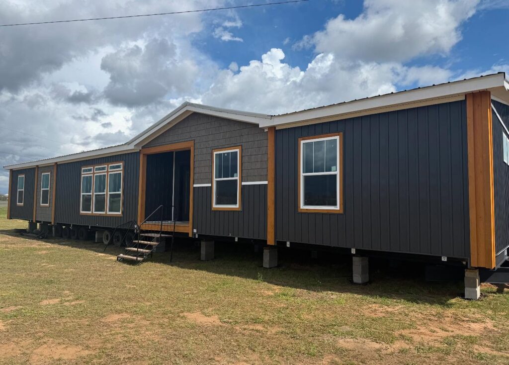A large, dark grey modular home stands elevated on stilts against a cloudy sky. The structure features multiple windows and wooden accents, evoking a modern and sturdy appearance.