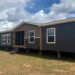 A large, dark grey modular home stands elevated on stilts against a cloudy sky. The structure features multiple windows and wooden accents, evoking a modern and sturdy appearance.