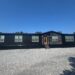 A modern black mobile home with multiple windows stands on a gravel lot under a clear blue sky. A small set of stairs leads to the front door.