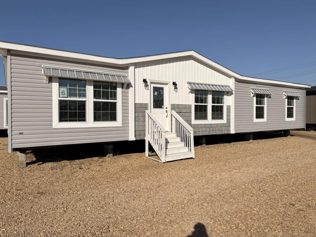 A modern gray manufactured home with white trim and striped window awnings stands on a gravel lot under a clear blue sky. It has steps leading to the door.