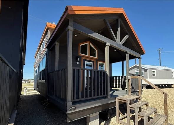 Cozy tiny house with a small porch and rustic wood steps, framed by a dark and warm color scheme. Bright blue sky creates a peaceful atmosphere.