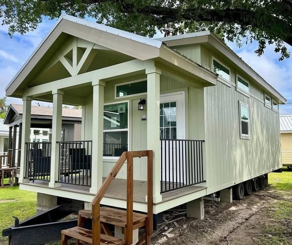 A small, light green tiny house sits on a grassy lot, featuring a welcoming front porch with black railings, wooden steps, and a gabled roof.