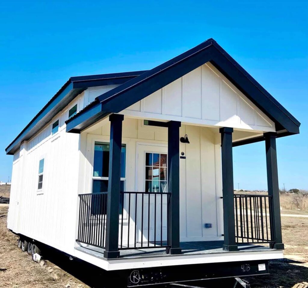 A small white tiny house with black trim stands under a clear blue sky. It features a covered front porch with railing and a glass-paneled door.