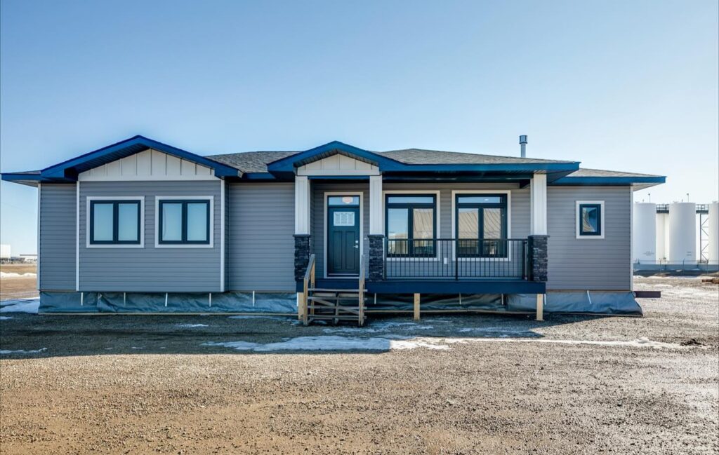 A modern modular home sits on a clear day, featuring gray siding, a dark roof, and a small porch with steps. The setting is open and expansive.