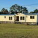 A single-story manufactured home with white siding and a blue roof sits in a grassy yard, surrounded by a black railing. Trees and a blue sky are in the background.