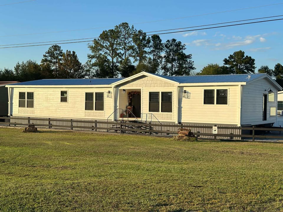 A single-story manufactured home with white siding and a blue roof sits in a grassy yard, surrounded by a black railing. Trees and a blue sky are in the background.