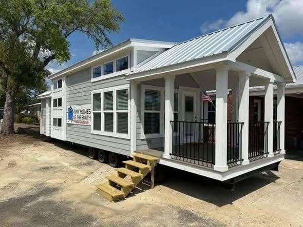 A modern tiny house on wheels with gray siding, white trim, and a small covered porch. A U.S. flag hangs outside. Bright, sunny day.