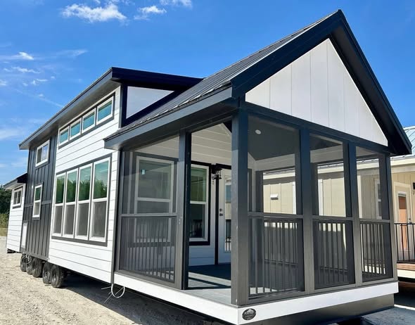 Modern tiny house on wheels with a gabled roof and gray-white exterior. Large windows and a screened porch create an airy, inviting vibe under a clear blue sky.
