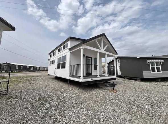 A modern tiny house with a gabled roof and white exterior sits on a gravel lot under a cloudy sky. It has large windows and a small front porch.
