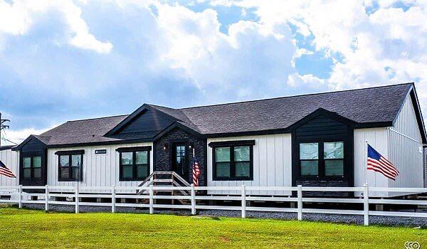Single-story black-and-white house with peaked roof, white fence, and American flags. Surrounded by lush green grass and under a cloudy blue sky.