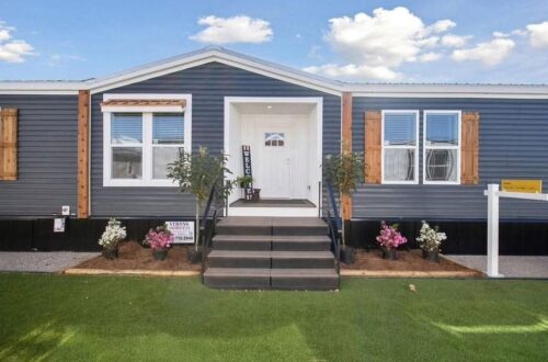 Single-story, blue modular home with white trim and wooden shutters, fronted by grass and flowers. The entrance is elevated with welcoming steps.