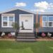 Single-story, blue modular home with white trim and wooden shutters, fronted by grass and flowers. The entrance is elevated with welcoming steps.