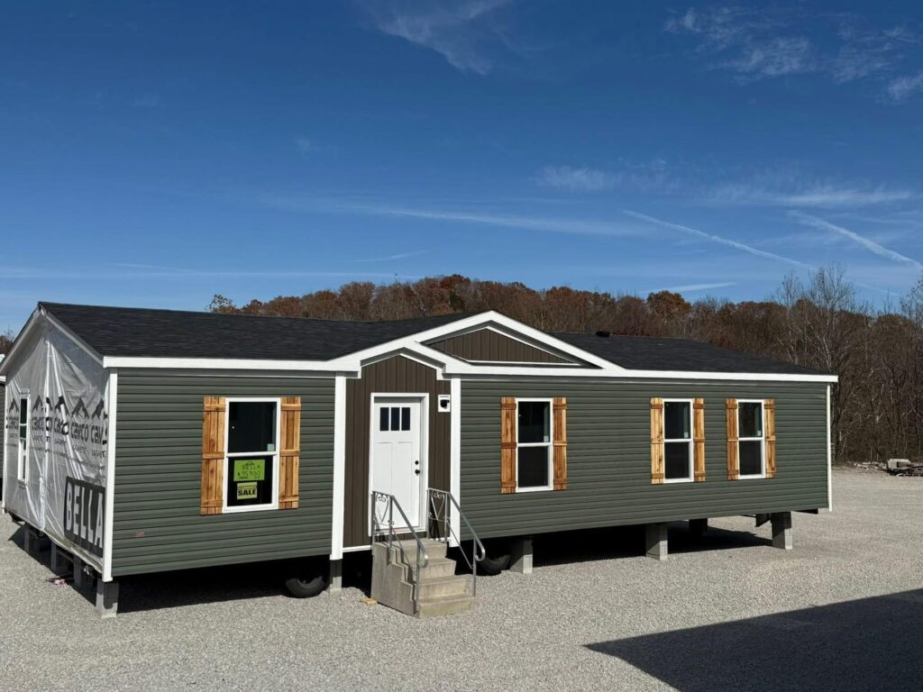 A green modular home with a white door and wooden shutters sits on a gravel lot under a clear blue sky, evoking a calm and inviting atmosphere.