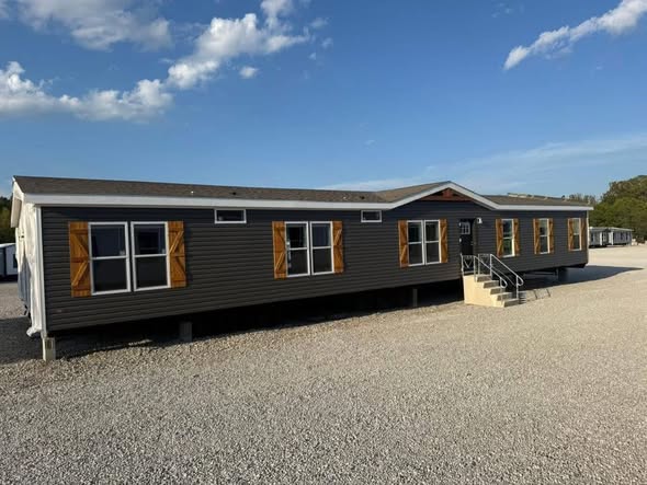 A long, dark gray modular home with orange shutters sits on gravel under a bright blue sky with scattered clouds. A small staircase leads to the entrance.