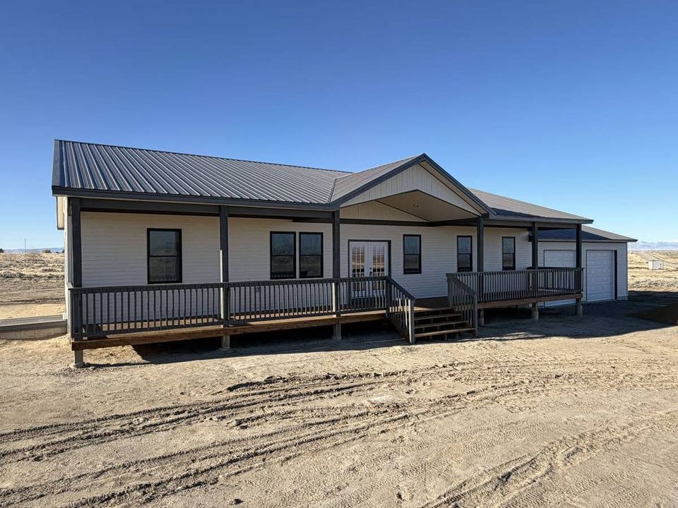 A modern one-story house with a metal roof and white siding, featuring a front porch with a wooden railing. The setting is a barren landscape under a clear blue sky.