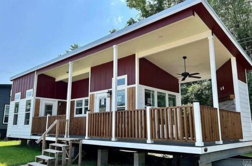 Modern tiny house with red and white exterior, elevated porch, wood railing, and ceiling fan against a clear blue sky and trees; cozy and inviting.