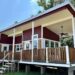 Modern tiny house with red and white exterior, elevated porch, wood railing, and ceiling fan against a clear blue sky and trees; cozy and inviting.