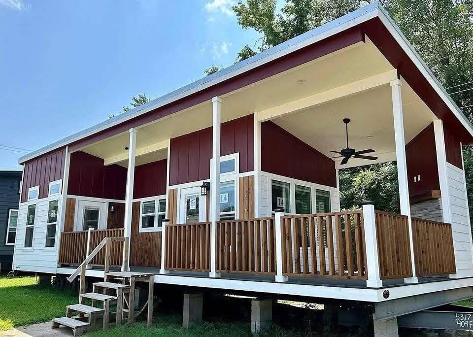 Modern tiny house with red and white exterior, elevated porch, wood railing, and ceiling fan against a clear blue sky and trees; cozy and inviting.