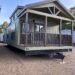 A green mobile home with a wooden porch, peaked roof, and black decorative door stands on a gravel lot under a clear blue sky.