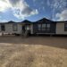 A large modular home with a blue and white exterior sits atop a gravel lot under a partly cloudy sky. Wooden steps lead to the front door.