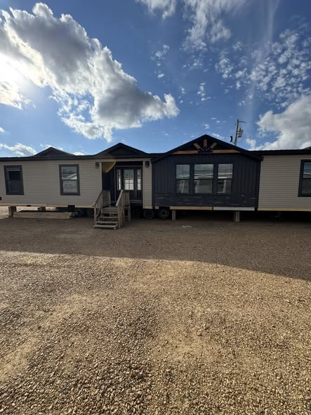 A large modular home with a blue and white exterior sits atop a gravel lot under a partly cloudy sky. Wooden steps lead to the front door.