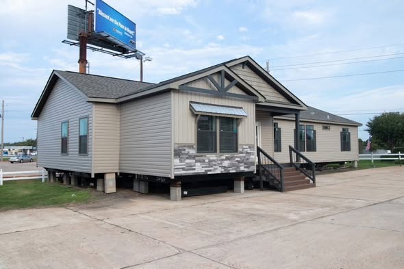 A modular home sits elevated on concrete blocks, with beige siding and stone accents. A small porch leads to its entrance. The sky is cloudy.