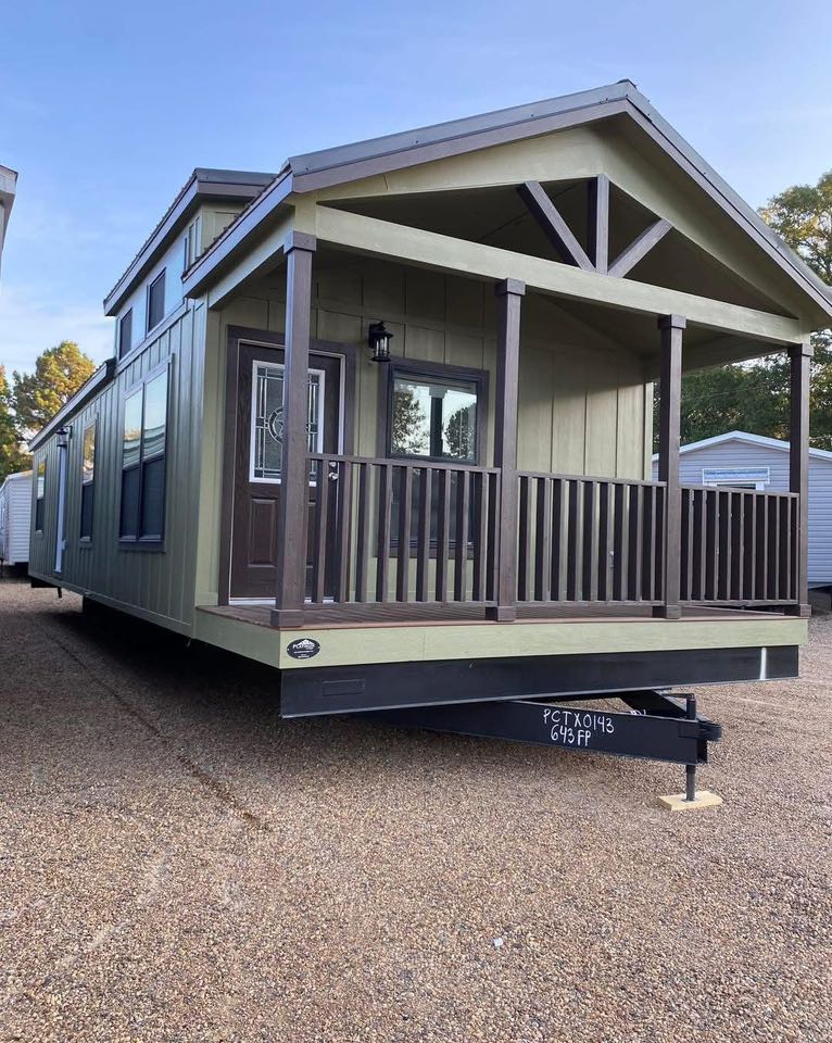 A small, mobile home with a green facade and wooden porch, set against a clear sky. The scene conveys simplicity and tranquility.