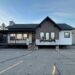 A modular home on a parking lot, elevated on blocks. It features wood accents, large windows, stone details, and a small porch. The sky is clear and blue.