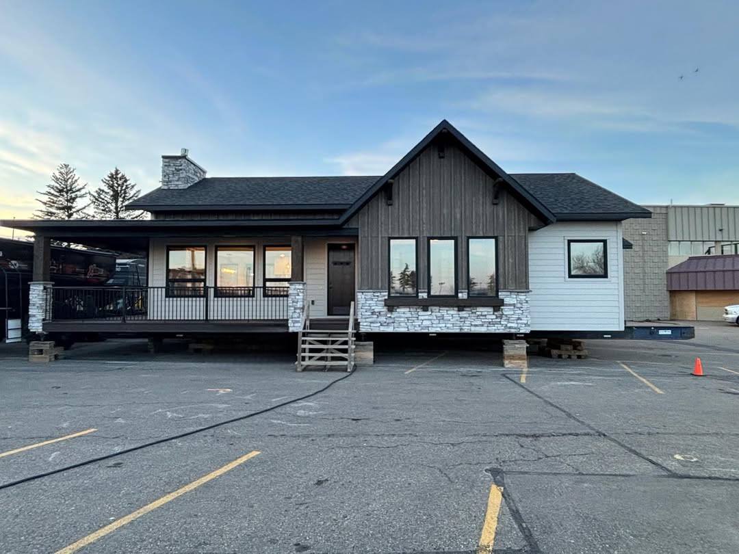 A modular home on a parking lot, elevated on blocks. It features wood accents, large windows, stone details, and a small porch. The sky is clear and blue.