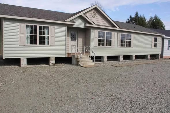 A light gray modular home on concrete blocks, set in a gravel area. Large windows and a small porch create a simple, functional appearance.