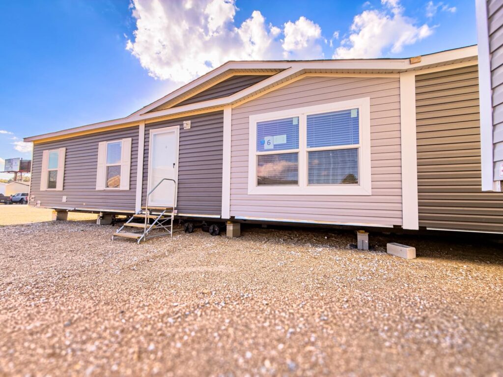 A manufactured home with beige and gray siding stands on a gravel lot under a bright blue sky with clouds. Steps lead to the white door, conveying a sense of openness.