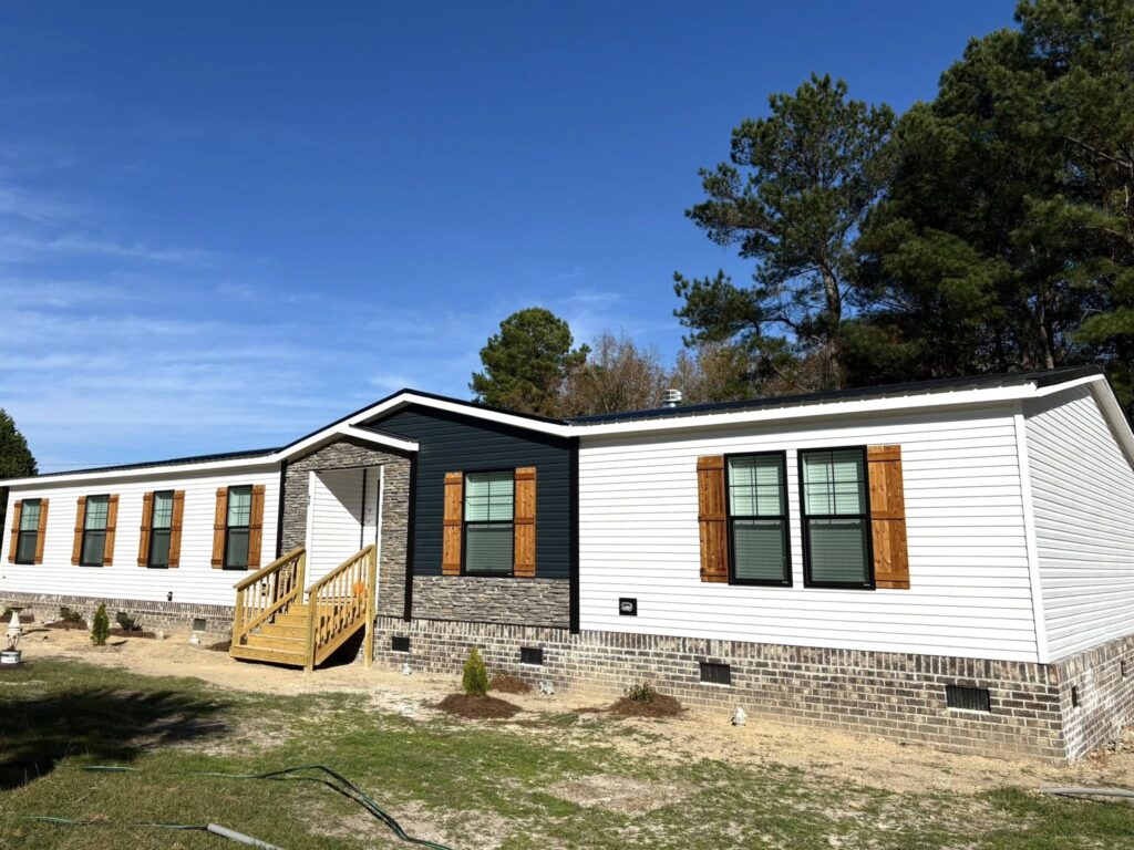 Modern, single-storey prefabricated home with white siding and brick base. Features wooden shutters, a small porch, and surrounded by trees under a clear blue sky.