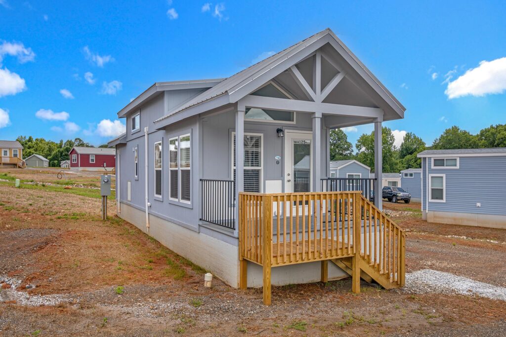 A modern tiny house with gray siding and a small wooden porch sits in a clearing under a bright blue sky. Other small homes are visible in the background.