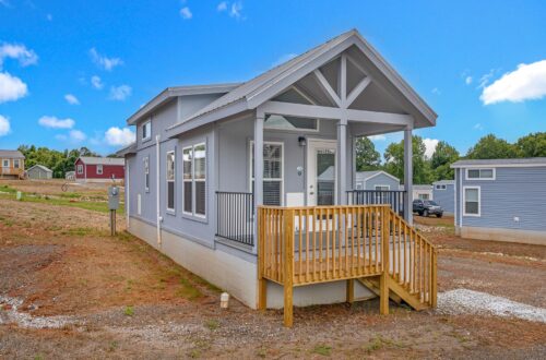 A modern tiny house with gray siding and a small wooden porch sits in a clearing under a bright blue sky. Other small homes are visible in the background.