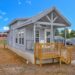 A modern tiny house with gray siding and a small wooden porch sits in a clearing under a bright blue sky. Other small homes are visible in the background.
