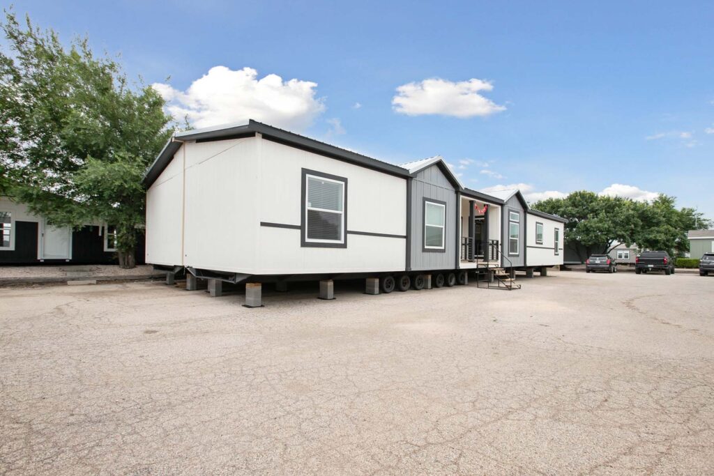 Three mobile homes sit side by side in a parking area, featuring modern design with white siding and dark trims. The sky is clear and blue.