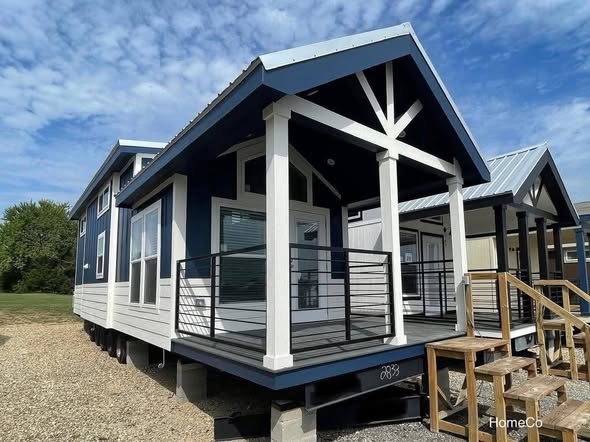 A modern tiny house with a blue and white exterior sits elevated on blocks. It features a small front porch with railings and stairs, under a clear blue sky.