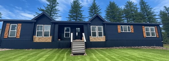 Blue house with wooden shutters, set against a backdrop of tall pine trees. The front features a small porch with stairs, conveying a peaceful, rustic feel.