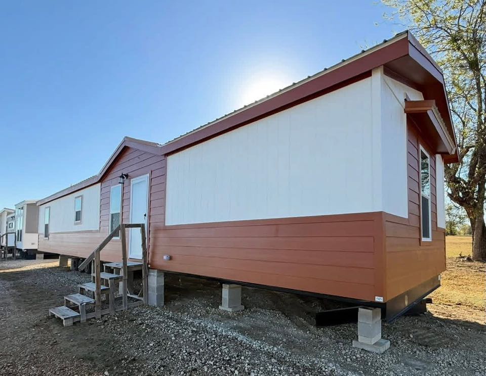 A mobile home with a red and white exterior sits elevated on blocks in a gravel lot. Wooden steps lead to the door. A tree and clear blue sky are visible.