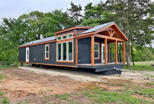 A modern tiny house with dark siding and wooden trim stands on a grassy plot. Large windows and a small porch create a cozy, inviting feel.